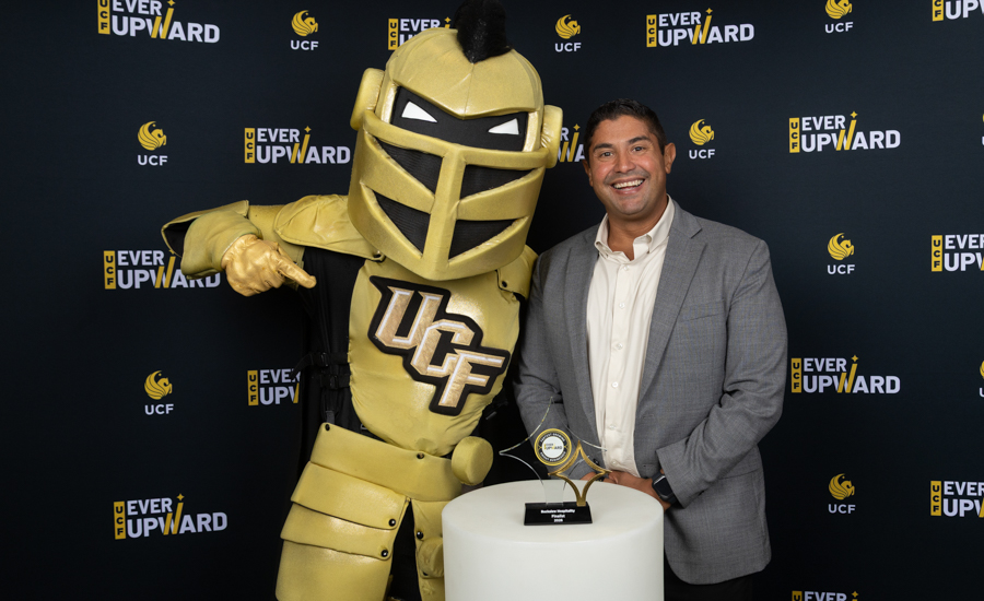 Man in a light gray suit posing with a gold knight mascot at a UCF 'Ever Upward' event, standing beside a white pedestal with a trophy; backdrop features UCF logos and 'Ever Upward' text.