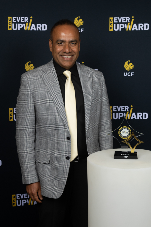Smiling man in a grey checkered blazer and pale yellow tie stands beside a trophy on a pedestal at an Ever Upward event backdrop.