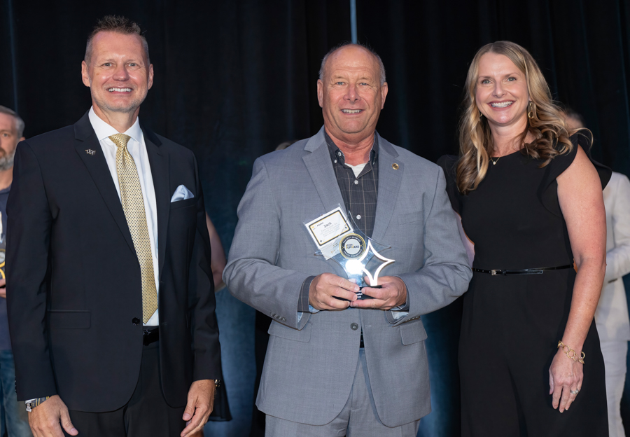 Three people on a stage posing; the man in the middle holds a glass award and certificate.