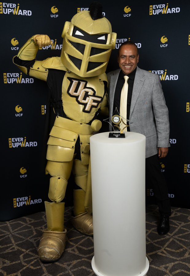 UCF knight mascot in gold armor flexes beside a man in a gray suit at an award event with a trophy on a pedestal behind them.