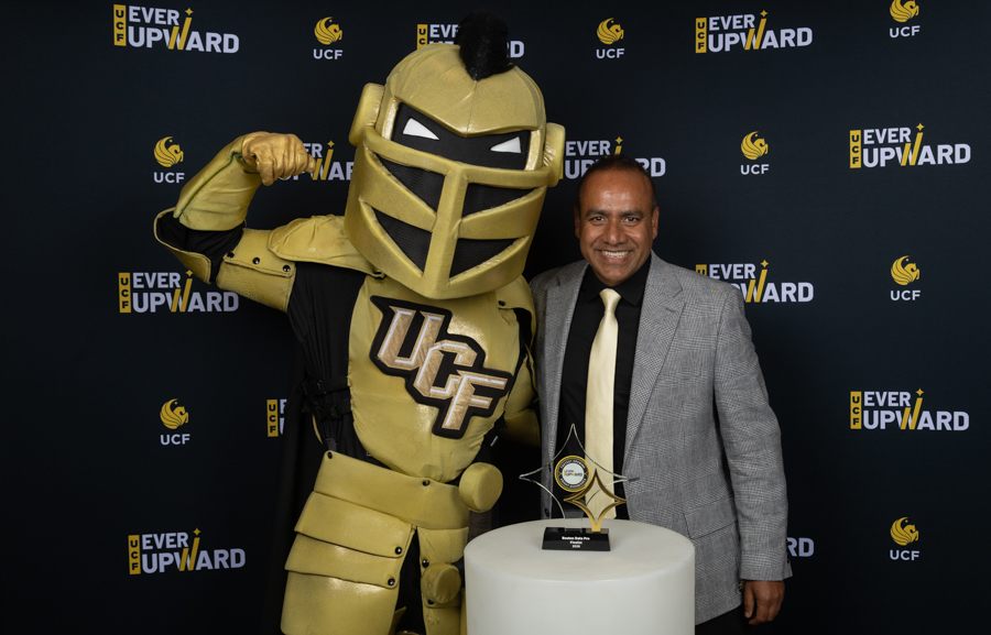 Golden UCF Knight mascot posing with a smiling man in a gray suit beside a trophy on a pedestal at an 'Ever Upward' event.