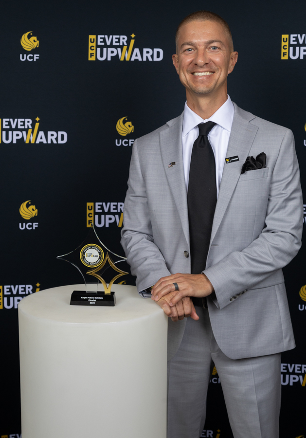 Man in a light gray suit and black tie smiles beside a trophy on a white pedestal at a UCF 'Ever Upward' event backdrop.