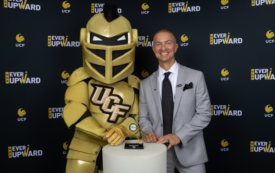 Golden Knight mascot next to a smiling man in a gray suit, posing with a trophy on a white pedestal at an UCF 'Ever Upward' event.