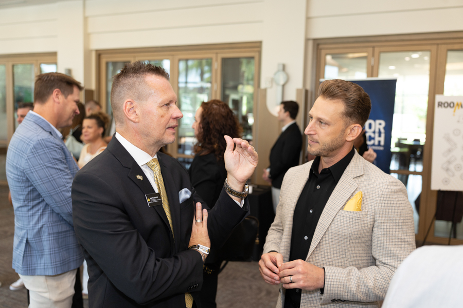 Two men in business attire talking at a networking event, with others chatting in the background.
