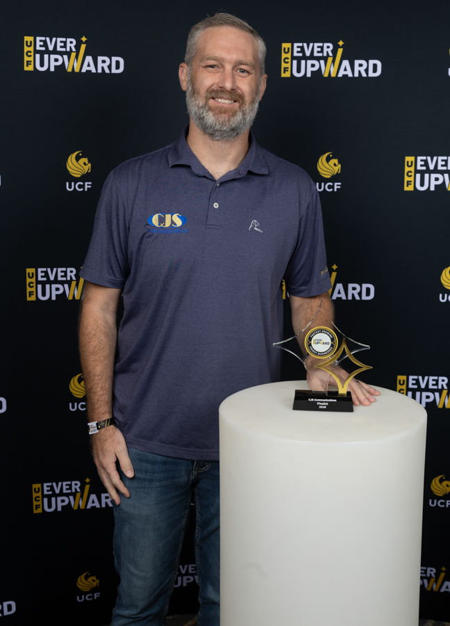 Smiling man with gray beard stands beside a white pedestal holding a glass trophy at a UCF Ever Upward event backdrop.
