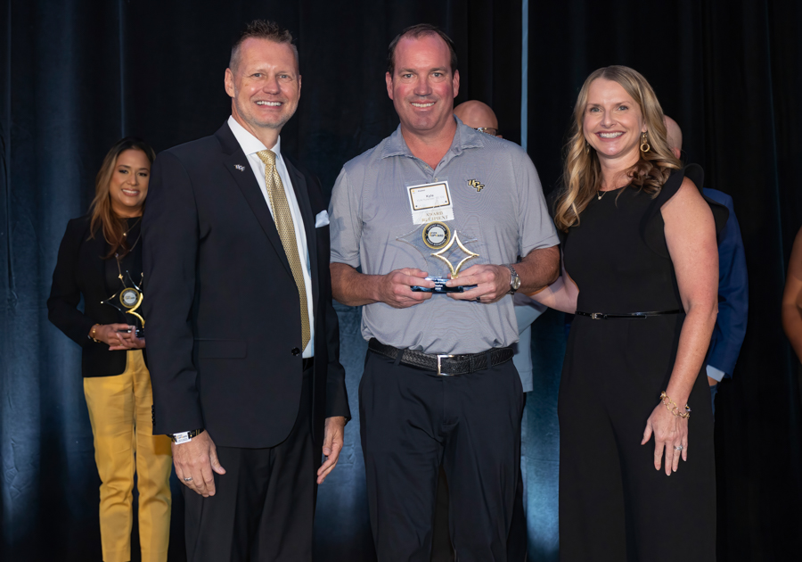 Man in gray polo receives an award on stage, flanked by a suited man and a woman in black dress.