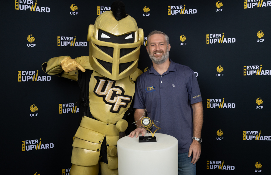 Man with beard posing with a gold UCF Knight mascot and a trophy at a UCF Ever Upward event.