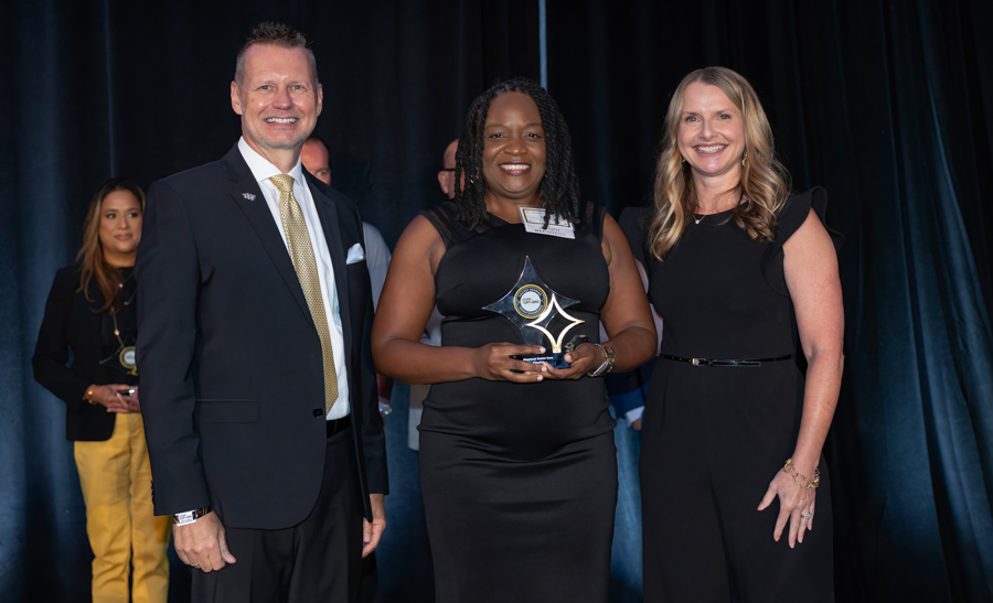 Three people on a stage for an awards event; center person holds a star-shaped trophy and smiles as others stand beside her in professional attire.