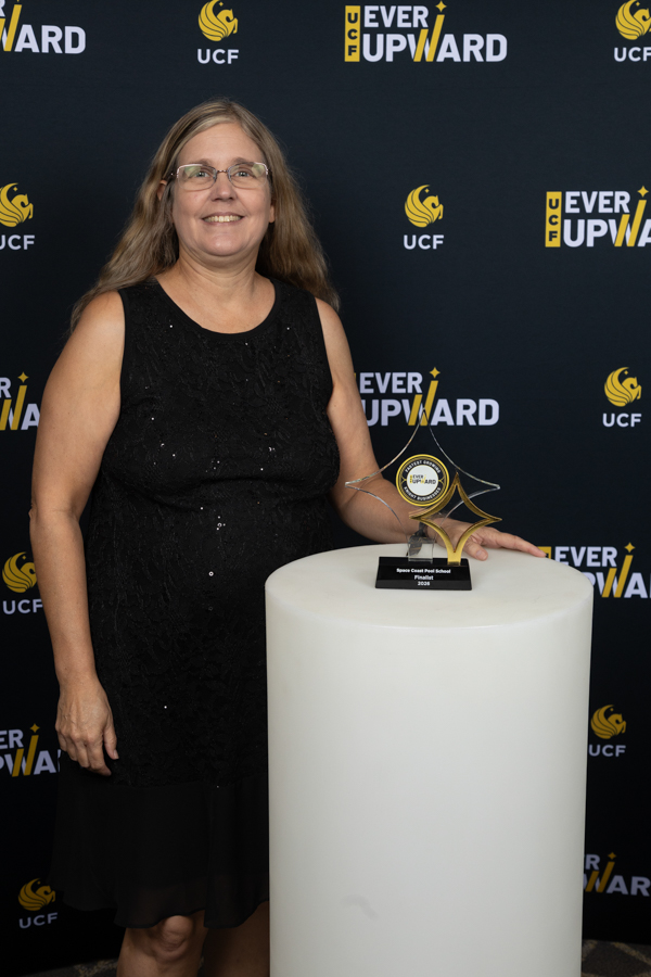 Smiling woman in a black sleeveless dress poses beside a white podium with a glass star trophy at a UCF Ever Upward event backdrop.