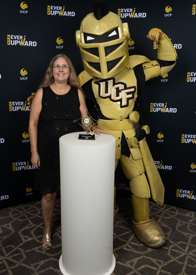 Smiling woman in a black dress poses with a gold UCF knight mascot and a trophy on a white pedestal.