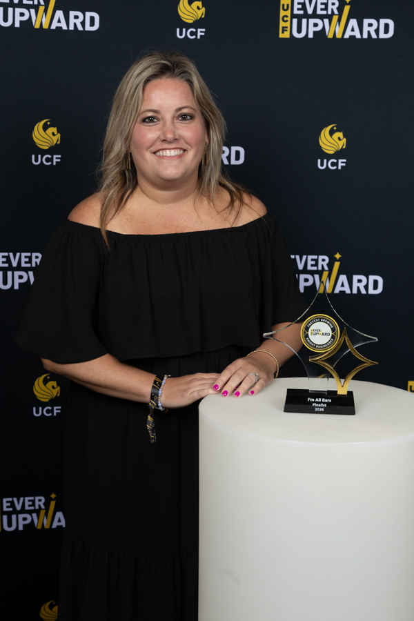 Smiling woman in a black off-the-shoulder dress stands beside a glass award on a white pedestal at a UCF event backdrop.