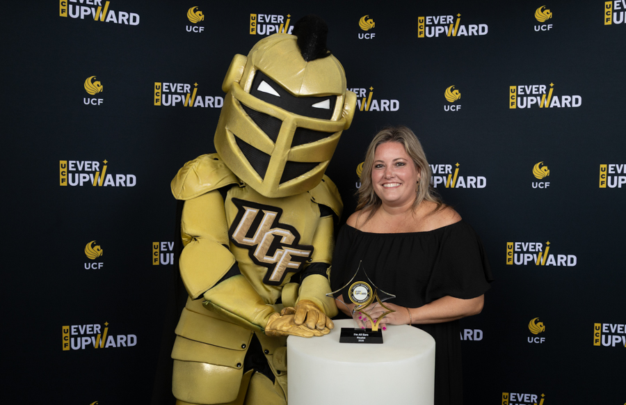 A gold UCF Knights mascot and a smiling woman in a black off-the-shoulder dress pose with a trophy on a white pedestal against a backdrop that says 'Ever Upward' and UCF logos.