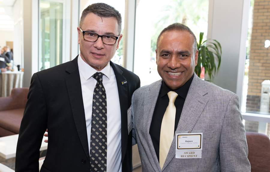 Two men in business attire posing together at an indoor event, one wearing a black suit and patterned tie, the other in a gray plaid jacket with a pale yellow tie and an award badge