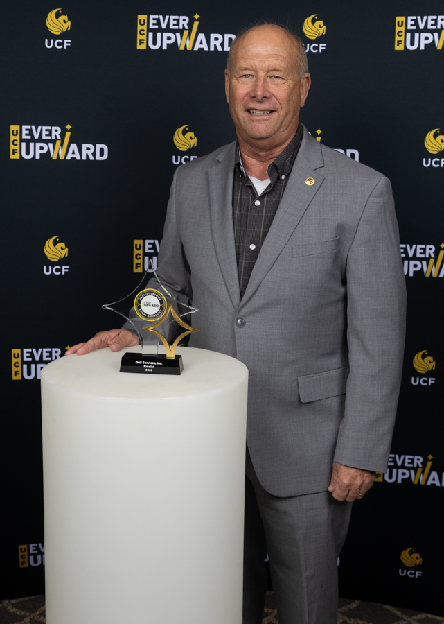 Man in a gray suit holds a star-shaped trophy on a white pedestal, posing at a UCF Ever Upward event backdrop.
