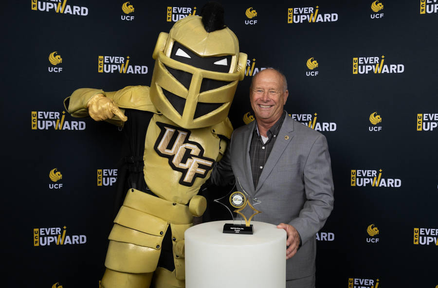 Smiling man in a gray suit posing with a gold UCF knight mascot at an 'Ever Upward' event backdrop.