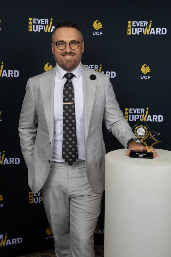 Smiling man in a light gray suit and patterned tie stands beside a trophy on a white pedestal at an Ever Upward event backdrop with UCF logos.