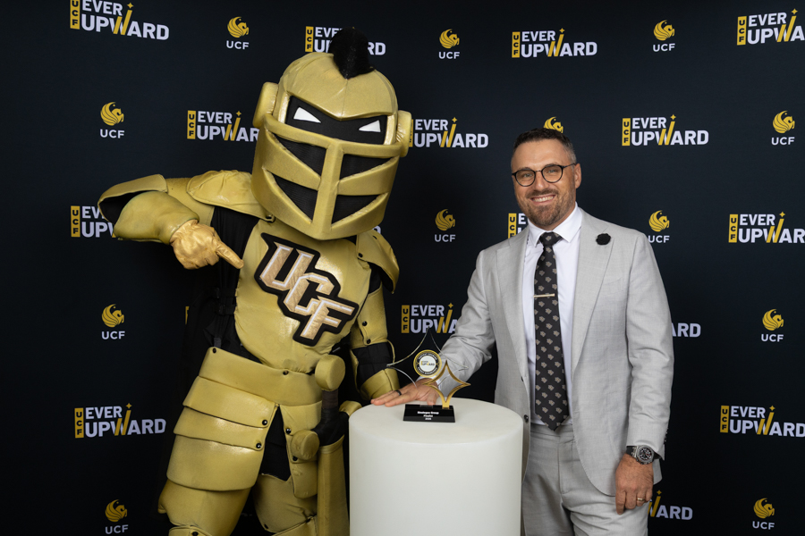 Gold-armored UCF mascot stands with a man in a gray suit, posing with a star-shaped award on a pedestal.