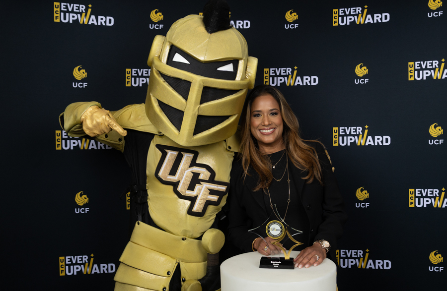 Woman posing with a gold UCF Knights mascot at an event backdrop reading 'Ever Upward'.