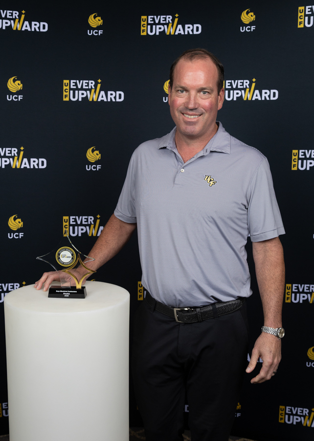 Smiling man in a light purple polo with a UCF emblem stands beside a glass trophy on a white pedestal at a UCF Ever Upward event backdrop.