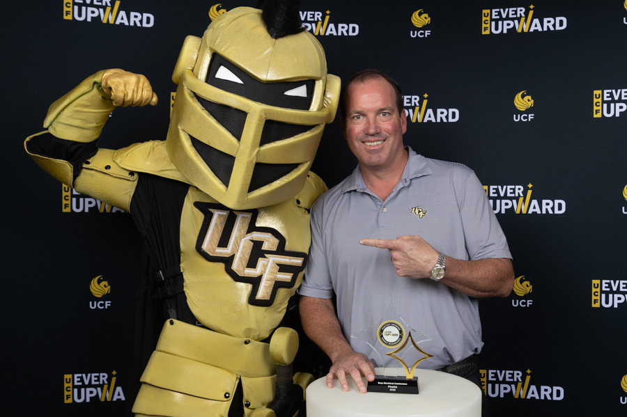 Man in gray polo posing with a gold knight mascot for UCF; the knight flexes a bicep while standing beside him in front of an Ever Upward backdrop, with a trophy on a pedestal between them.