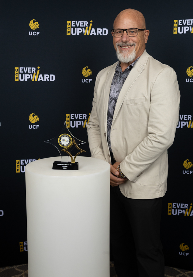 Smiling bald man with glasses in a light blazer posing with a glass star trophy on a white pedestal at an UCF Ever Upward event backdrop.