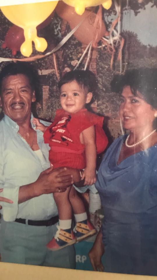 Family of three at a birthday party: man in light blue shirt holding a toddler in red outfit, with a woman in a blue dress beside them and yellow balloons overhead.