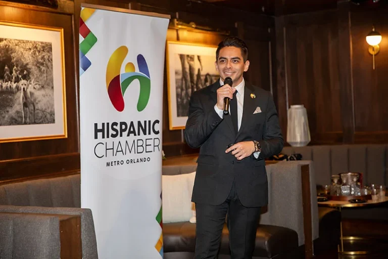 Man in a dark suit speaking into a microphone at a Hispanic Chamber event, with a banner reading 'Hispanic Chamber Metro Orlando' beside him.