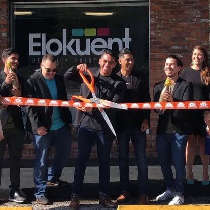 Seven people stand outside a brick building as the central figure cuts an orange ribbon in front of a sign that reads Elokuent, celebrating a storefront opening.