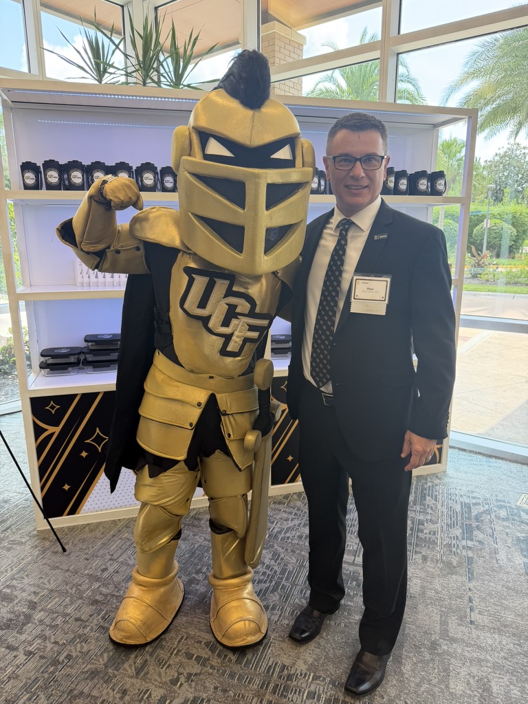 Man in a suit posing with a gold knight mascot featuring a UCF logo indoors at an event venue.