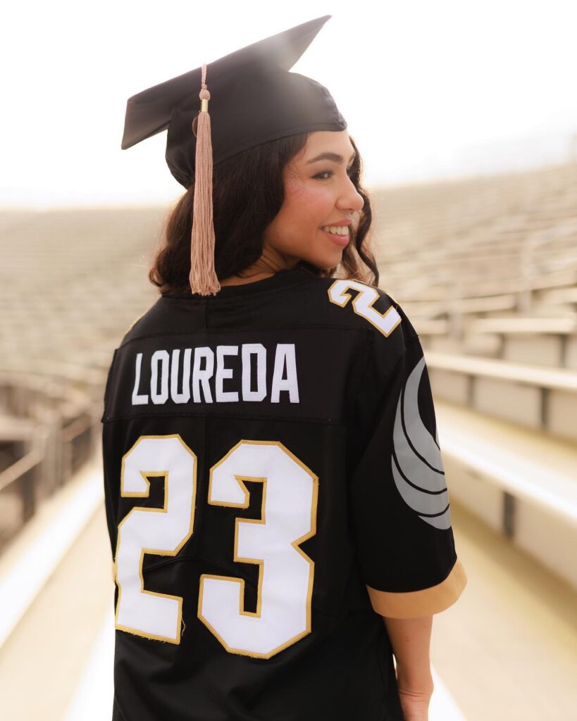 Smiling graduate in a black cap and gown, seen from behind on bleachers; jersey reads LOUREDA and number 23.