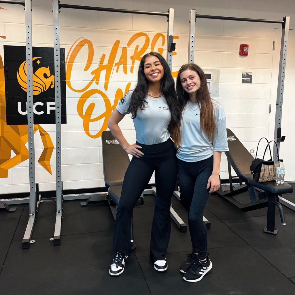 Two women pose together in a gym, smiling, wearing light blue shirts and dark athletic pants with workout equipment in the background.