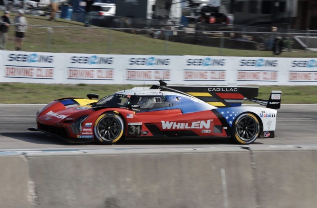 Colorful race car number 31 with Whelen branding speeding along a race track, Cadillac on the rear wing, blue-red-white star livery on the body.