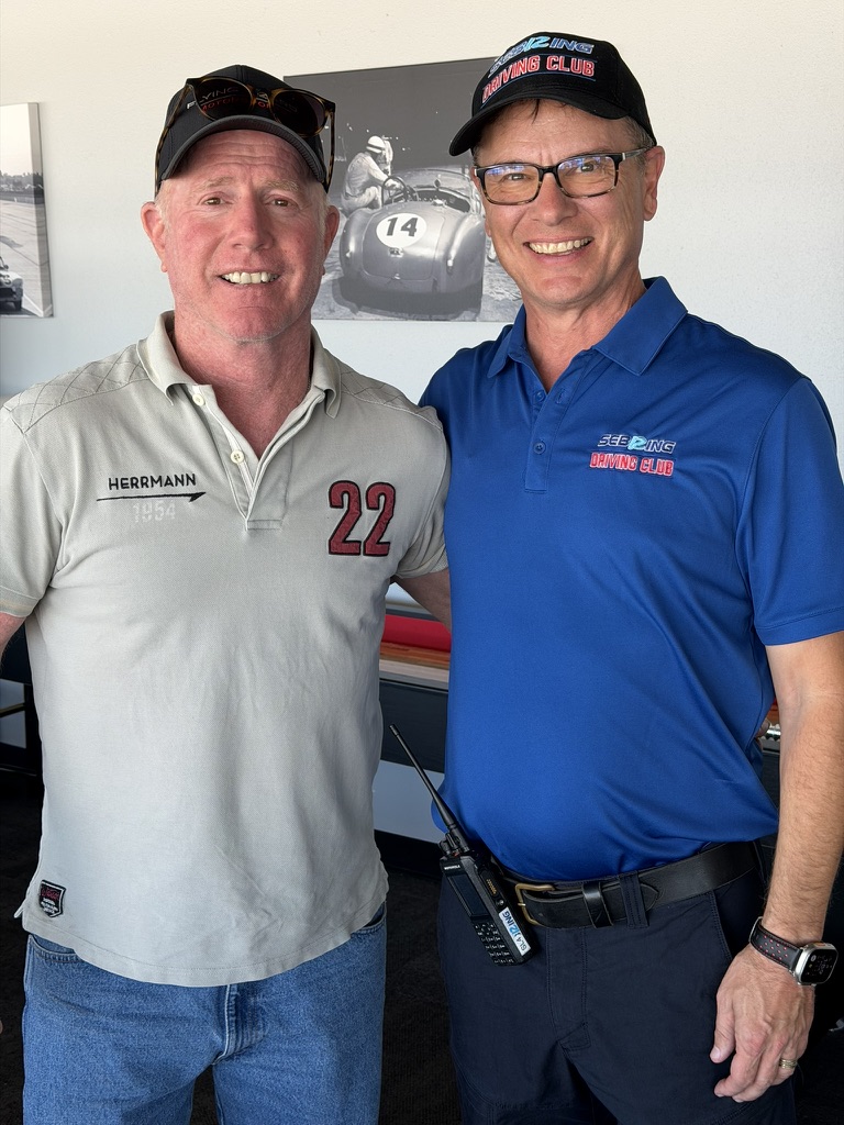Two men posing together at a race club; one wears a gray polo with the number 22 and 'Herrmann', the other in a blue polo with the club logo, both smiling.