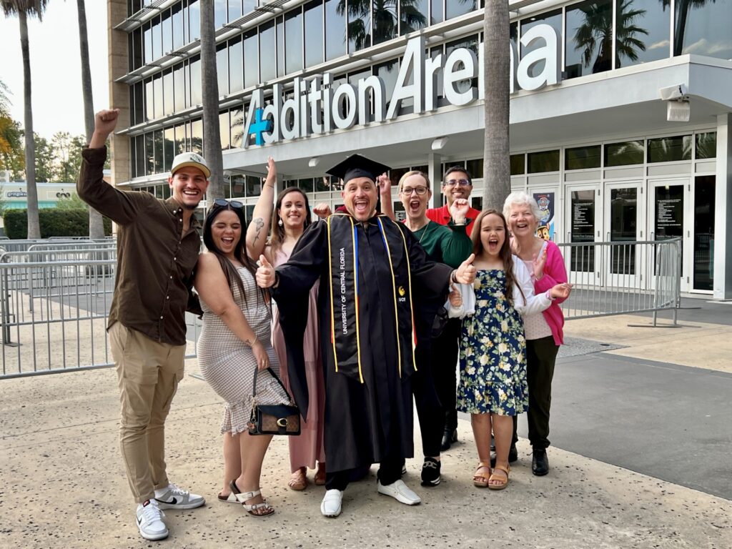 Graduate in cap and gown poses with smiling family and friends outside a modern building with large lettering in the background, cheering.