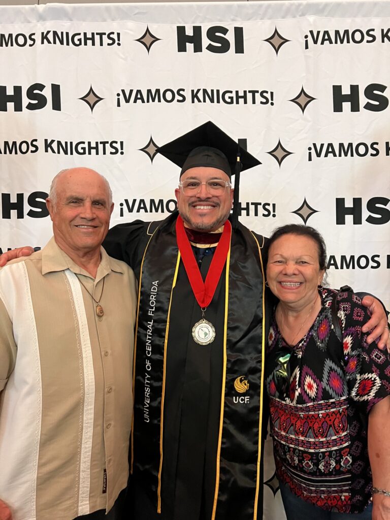 Graduate in cap and gown with medal, flanked by two relatives, posing in front of an HSI banner reading ¡Vamos Knights!