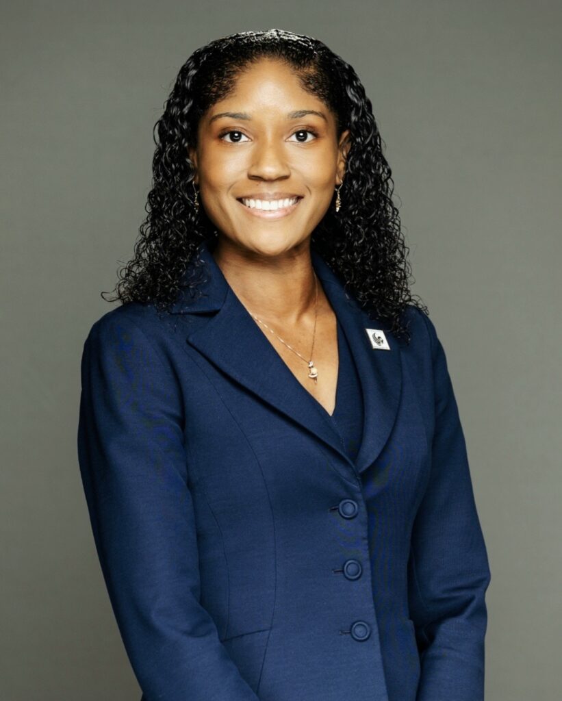 Professional woman in a navy blazer smiling at the camera, badge on the lapel, neutral gray backdrop.