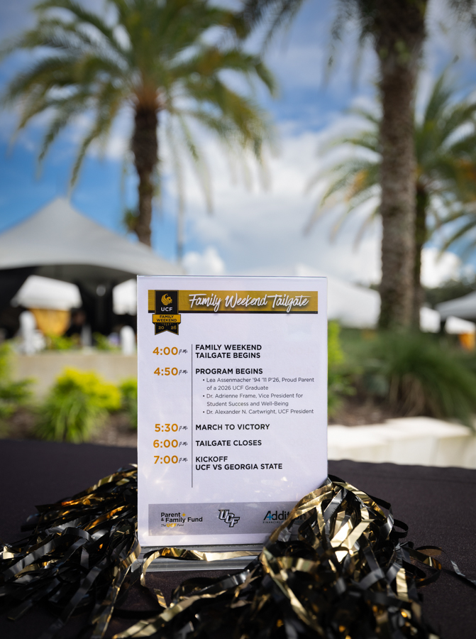 Printed event schedule for 'Family Weekend Tailgate' on a table with black-and-gold streamers; outdoor venue with palm trees in background.