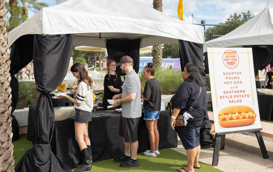 People wait in line at a food booth under white tents during an outdoor event; a sign advertises a hot dog with Southern-style potato salad nearby.