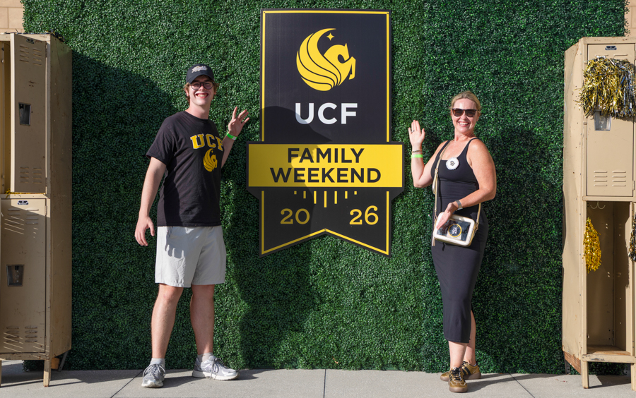 Two people pose on either side of a UCF Family Weekend sign against a green hedge backdrop.