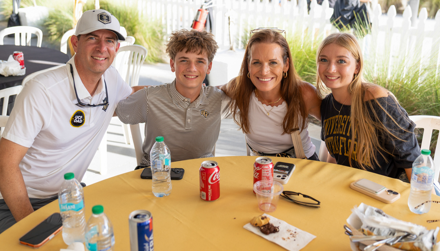 Four people sit at a yellow outdoor table, smiling for the photo, with drinks and a snack plate in front of them.