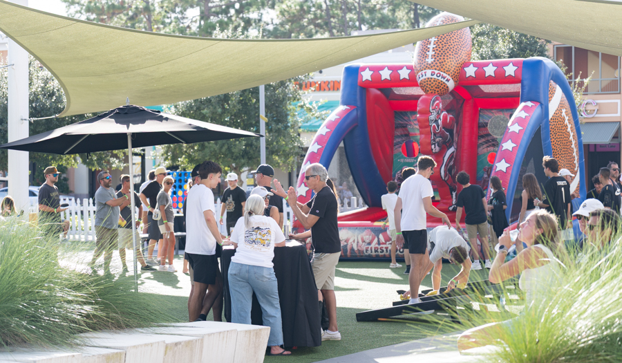 Outdoor festival scene with a large football-themed inflatable obstacle course in the background and people gathered around a high table under shade sails.