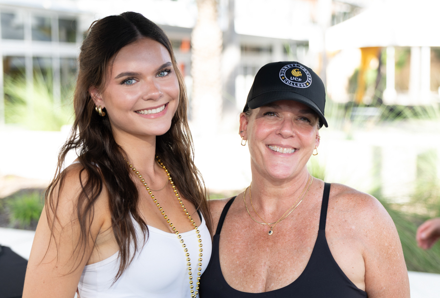 Two smiling women posing outdoors; a younger woman in a white top with a gold bead necklace and an older woman in a black tank top and cap beside her.