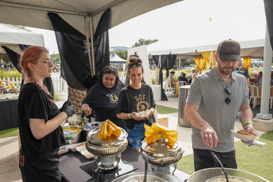 People serving food at an outdoor event under tents, with chafing dishes and yellow napkins on the serving table