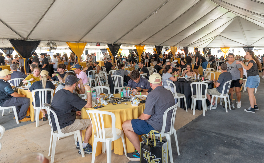 Crowded outdoor banquet under a large tent, people dining at round tables with yellow tablecloths.