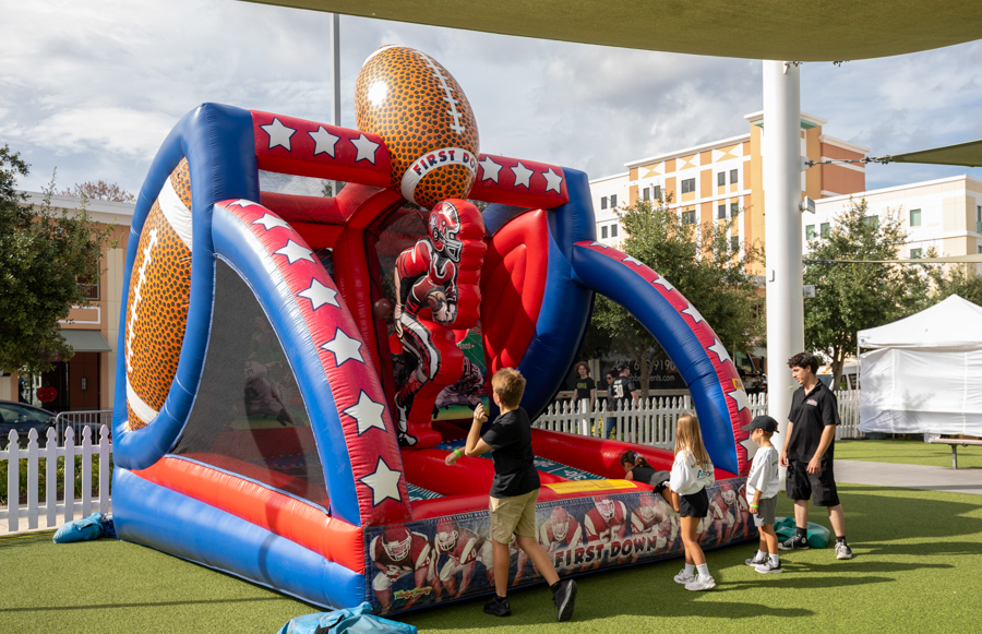 Colorful inflatable football-themed bounce house with kids lining up to enter at an outdoor event in the sun.