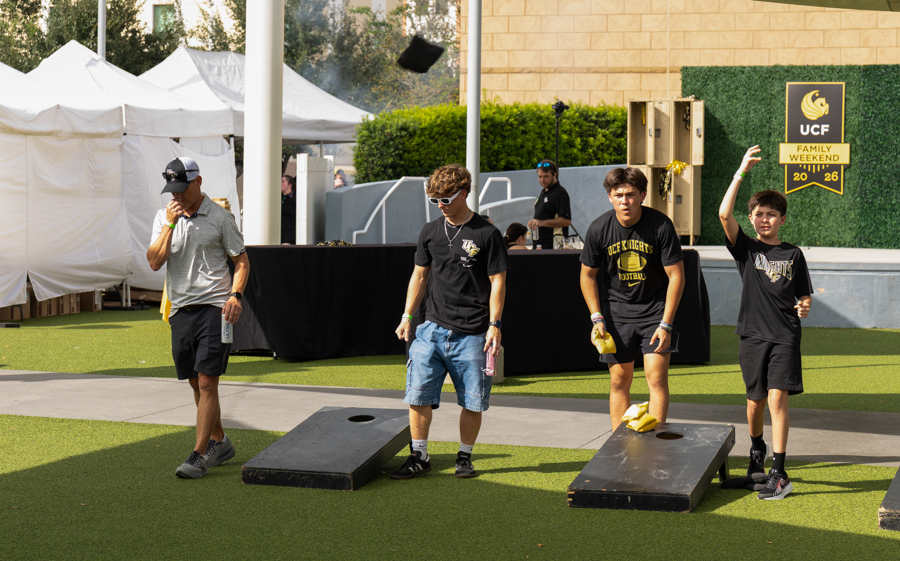 Group of four men playing a bean bag toss (cornhole) game outdoors during USC Family Weekend, lining up shots toward holes on two boards with a tent backdrop.