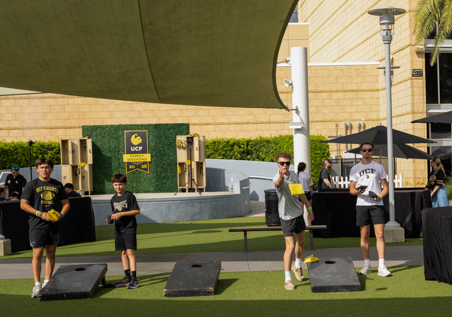 Group of college students playing cornhole on a sunny UC campus setup with tents and a stage in the background.