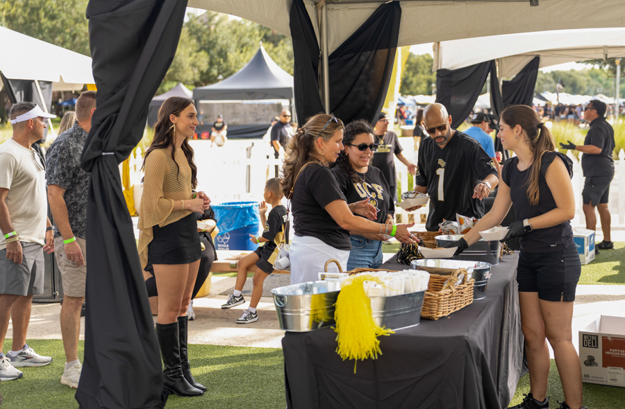Group of people at an outdoor festival near a food serving table under a canopy, volunteers handing out plates.