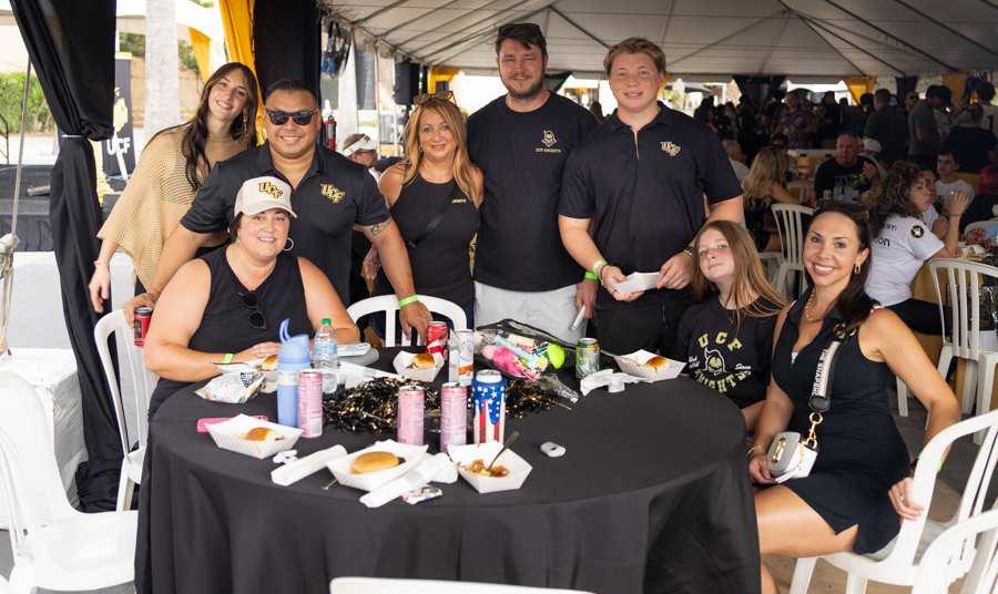 Group of eight people posing around a round table under a large tent at an outdoor event, smiling for the camera with food and drinks on the table.