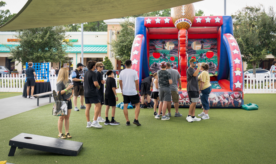 Group of people waiting in line at a bright inflatable basketball game booth outdoors on a sunny day, with a white picket fence nearby.